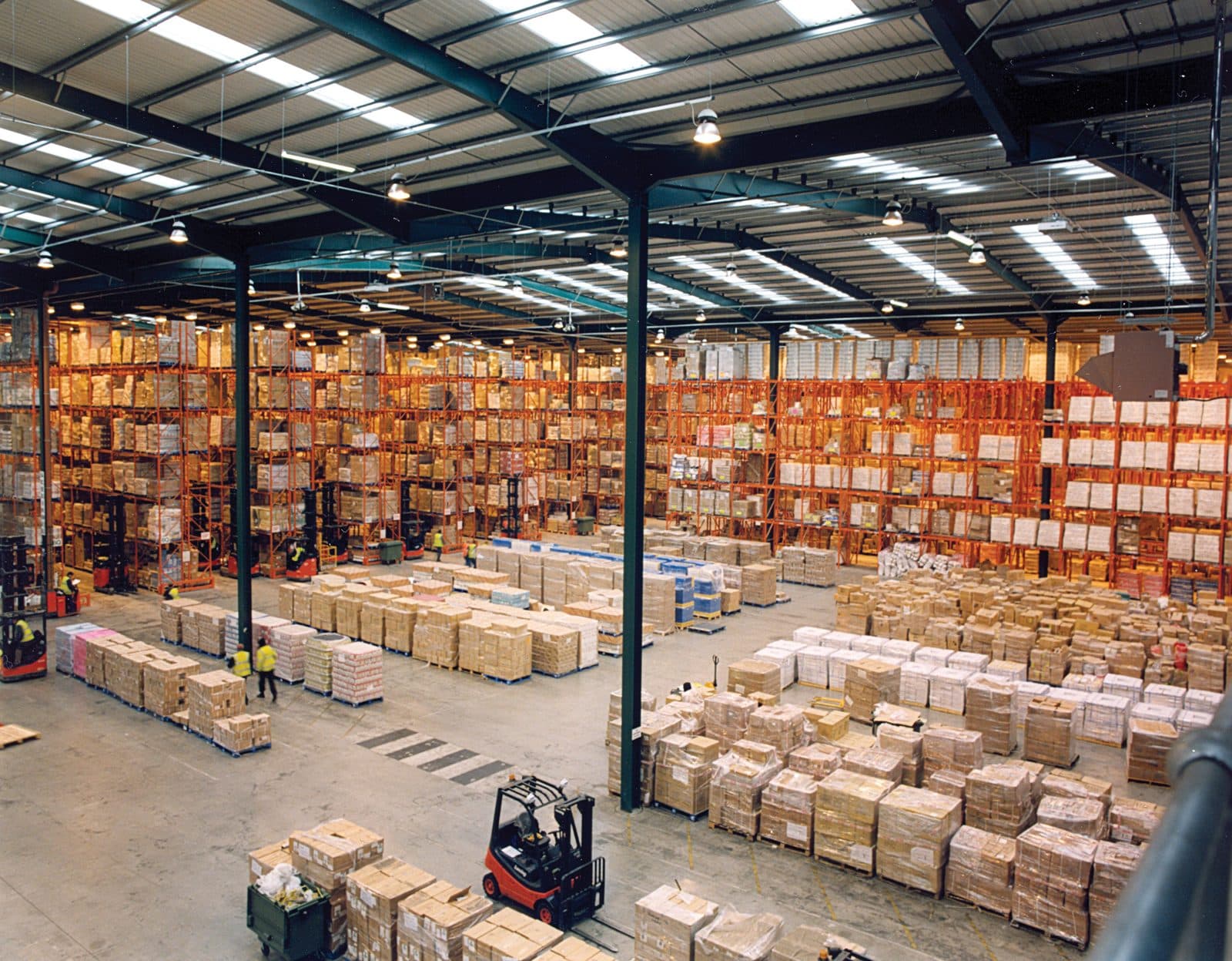 Interior of a modern warehouse showing tall metal pallet racking filled with palletized goods and a forklift in the aisle, with no people in frame.